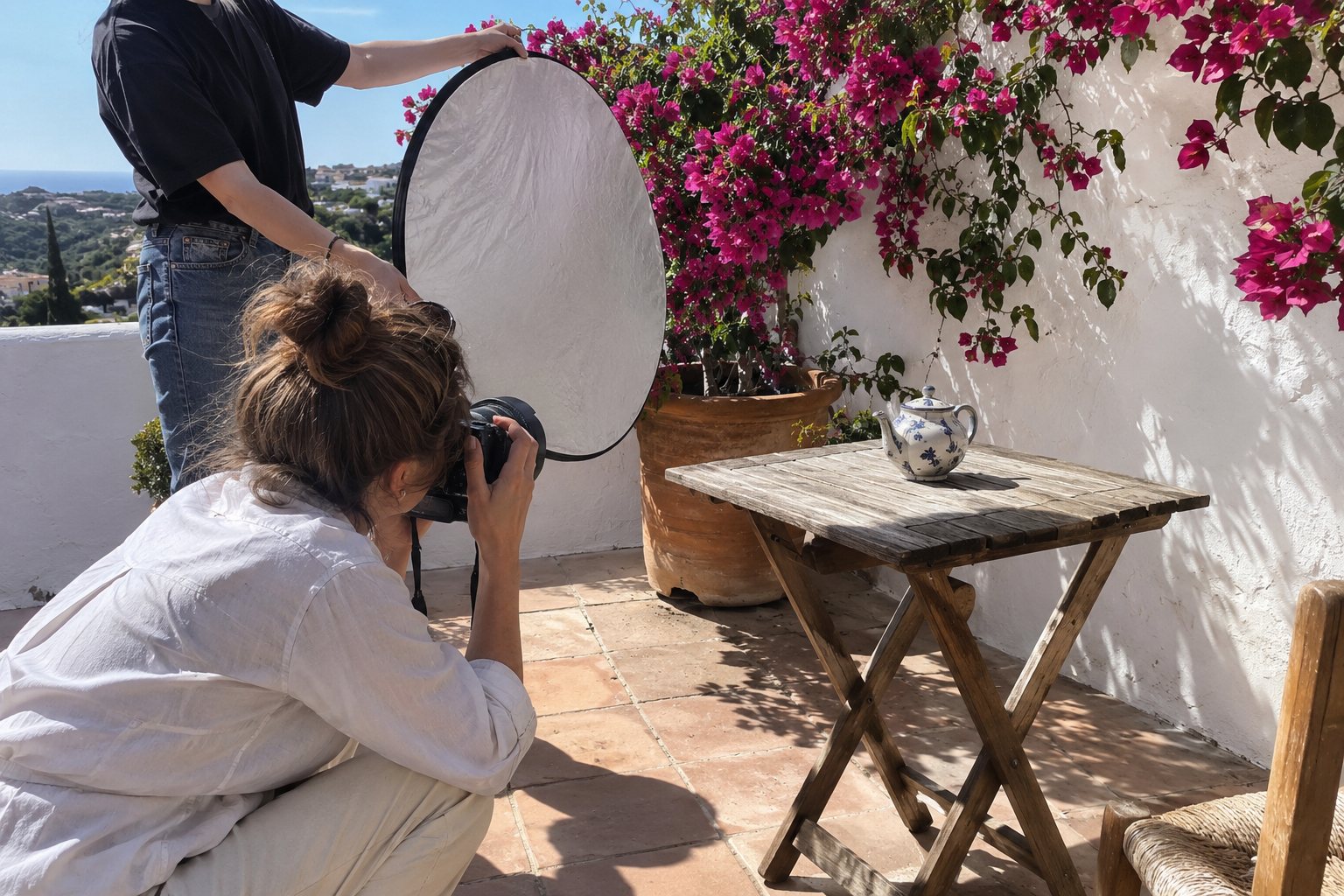 Een fotografe hurkt op een zonnig terras met terracotta tegels en bougainville om een theepot te fotograferen, een assistent houdt een reflectiescherm vast