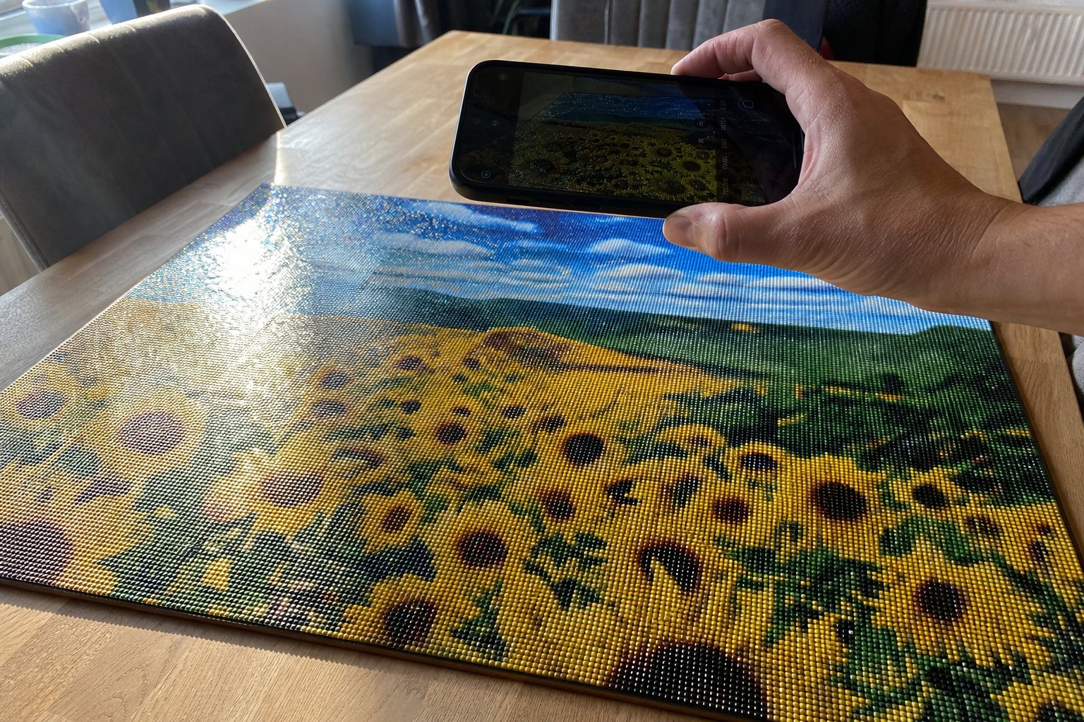 Een afgemaakte diamond painting van een zonnebloemenveld op tafel, met een telefoon erboven en een felle glans van het raamlicht op de steentjes